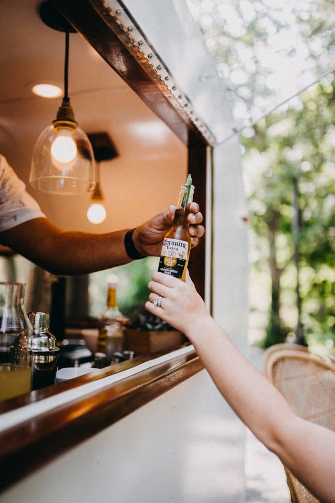 A close-up of a hand receiving a beer bottle through a food truck window outdoors.